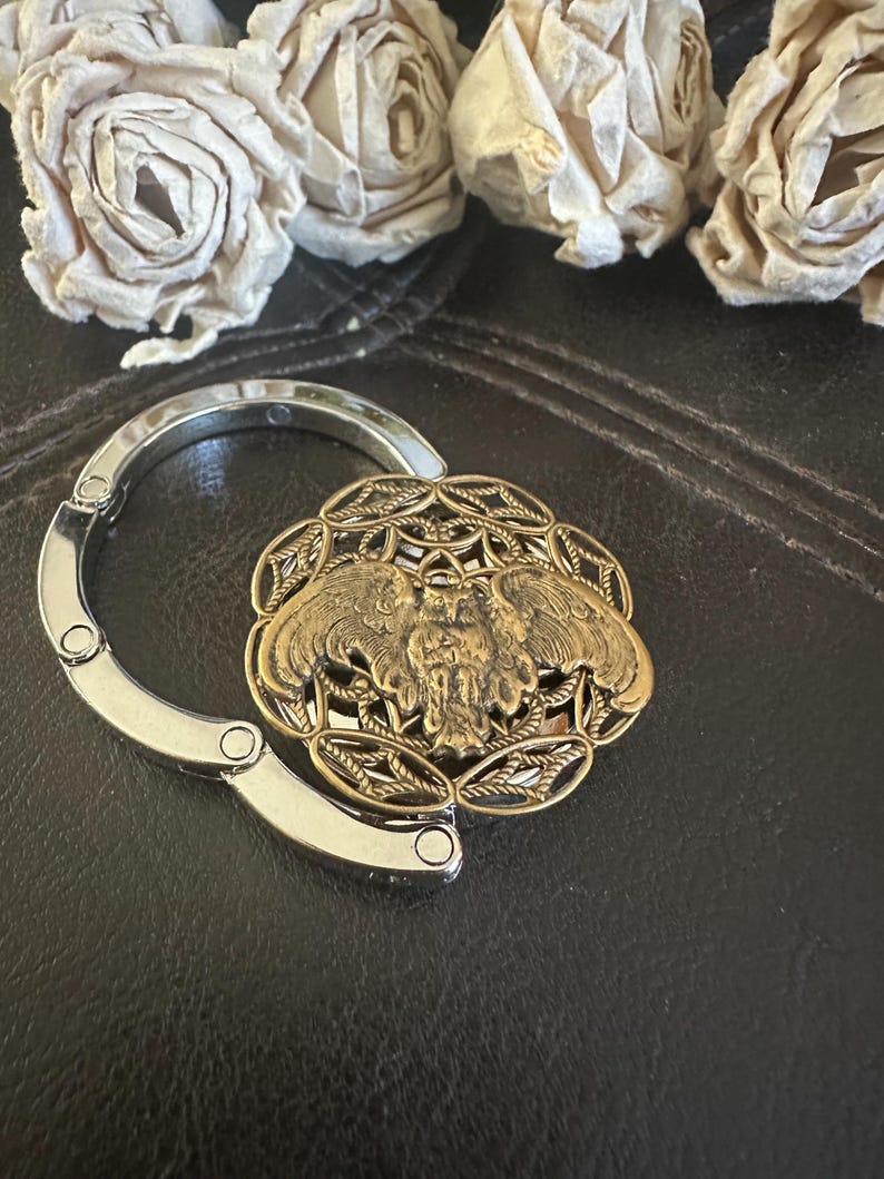May include: A circular metal bag hanger featuring a bronze-toned owl emblem centered within an intricate filigree pattern. The hanger has a polished silver-colored folding hook mechanism that extends from the side of the decorative owl centerpiece.