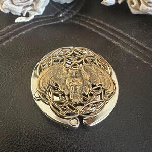May include: A round, foldable metal purse hook featuring a brass-toned owl emblem centered on a decorative, openwork filigree background. The base of the hook is polished silver-tone metal, designed to hang a bag from a flat surface edge.