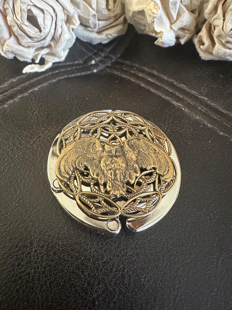May include: A round, foldable metal purse hook featuring a brass-toned owl emblem centered on a decorative, openwork filigree background. The base of the hook is polished silver-tone metal, designed to hang a bag from a flat surface edge.