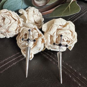 May include: A pair of silver-toned earrings with a dagger design. Each earring features a cross-shaped top adorned with black gemstones and a long, pointed spike. The earrings are displayed with cream-colored fabric roses.