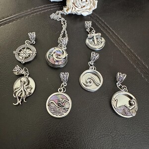 May include: A collection of silver-toned pendants with nautical themes. Designs include a compass, octopus, anchor, waves, and a mermaid. One pendant features a colorful, iridescent inlay. The pendants are displayed on a dark, textured surface.