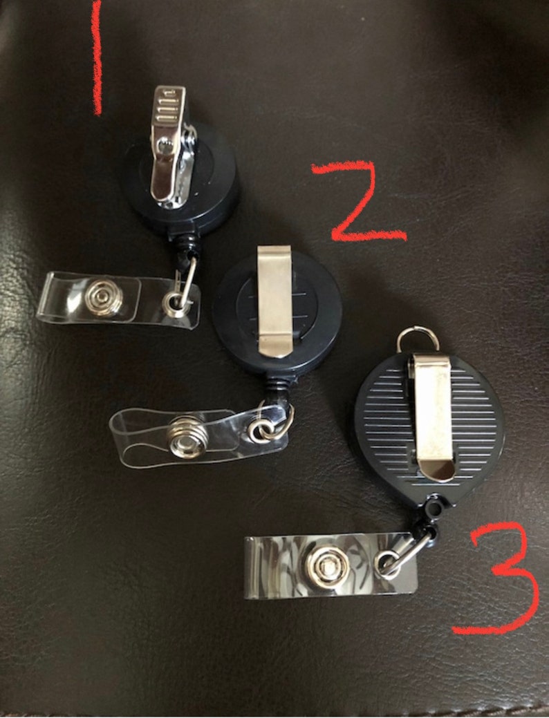 K&ouml;nnte beinhalten: Drei schwarze, einziehbare Ausweishalter mit durchsichtigen Plastikb&auml;ndern. Jeder Halter hat einen silbernen Clip und einen silbernen Drehhaken.
