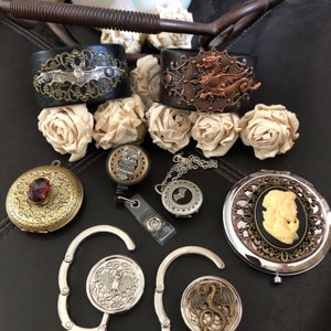 May include: Two leather cuffs featuring silver bat and copper dragon filigree, a round brass locket with a red stone, an owl badge reel, a silver bat locket, a cameo compact mirror, and two silver purse hooks with bat and dragon motifs.