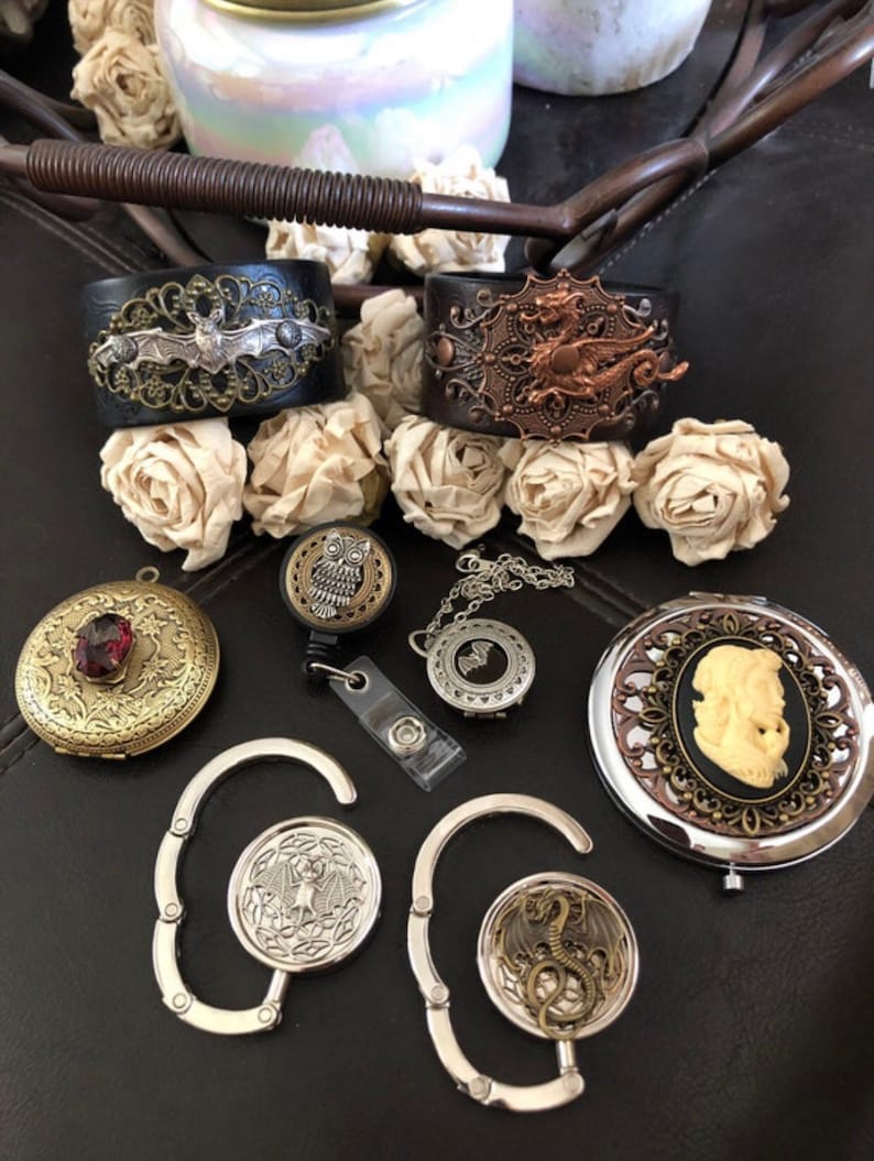 May include: Two leather cuffs featuring silver bat and copper dragon filigree, a round brass locket with a red stone, an owl badge reel, a silver bat locket, a cameo compact mirror, and two silver purse hooks with bat and dragon motifs.