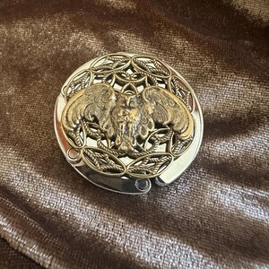 May include: A round, silver-toned metal bag hanger featuring a central, raised brass-colored owl with outstretched wings. The owl is surrounded by an intricate, openwork filigree pattern of interconnected leaves and vines within the circular frame.