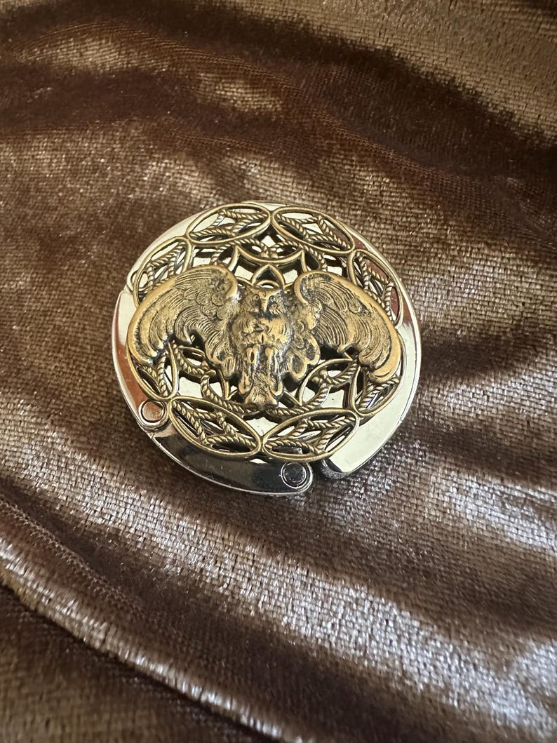 May include: A round, silver-toned metal bag hanger featuring a central, raised brass-colored owl with outstretched wings. The owl is surrounded by an intricate, openwork filigree pattern of interconnected leaves and vines within the circular frame.
