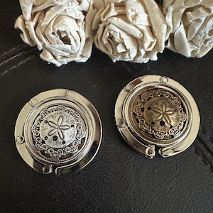 May include: Two circular metal purse hooks featuring a sand dollar design in the center. One hook has a bright silver finish, while the other has an antique bronze finish. Both items have a smooth, polished outer ring and a decorative floral border.