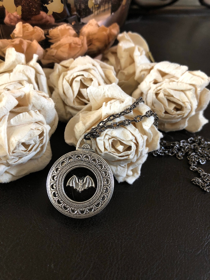 May include: A silver necklace with a circular pendant featuring a detailed bat design. The pendant has a black background and a silver bat in the center. The necklace is on a black surface with white flowers.
