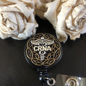 K&ouml;nnte beinhalten: Schwarzer einziehbarer Ausweishalter mit einem goldenen und silbernen medizinischen Symbol und dem Text "CRNA".