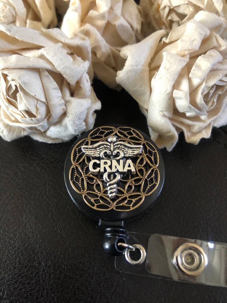K&ouml;nnte beinhalten: Schwarzer einziehbarer Ausweishalter mit einem goldenen und silbernen medizinischen Symbol und dem Text "CRNA".