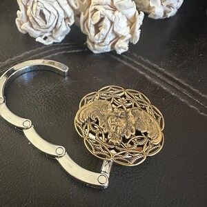 May include: A silver-toned metal folding bag hanger featuring a circular decorative top. The top piece has an antique gold finish with an intricate openwork border and a central relief design of an owl with wings spread wide in flight.