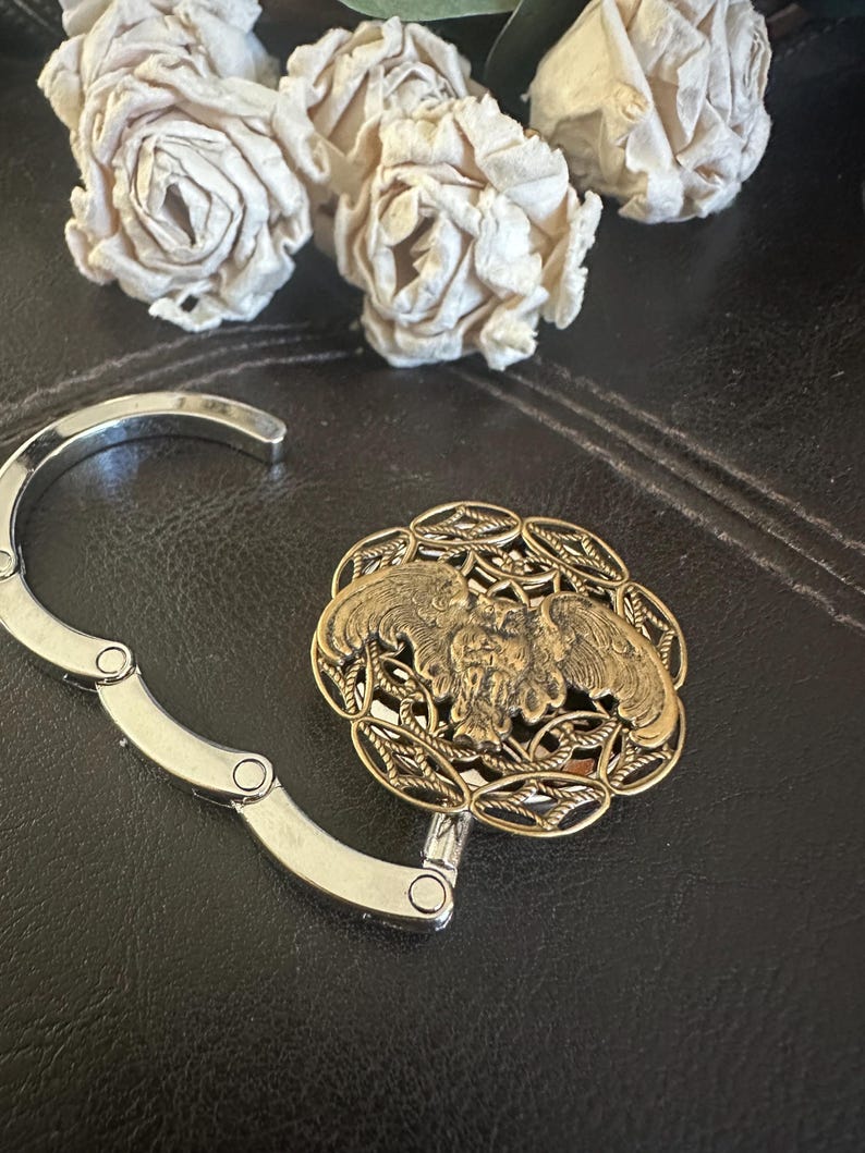 May include: A silver-toned metal folding bag hanger featuring a circular decorative top. The top piece has an antique gold finish with an intricate openwork border and a central relief design of an owl with wings spread wide in flight.
