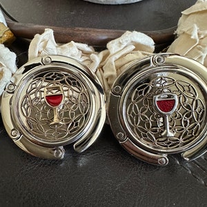 May include: Two silver-colored purse hooks with a wine glass design. Each hook has a round, decorative top with a red wine glass in the center. The hooks are on a black surface.