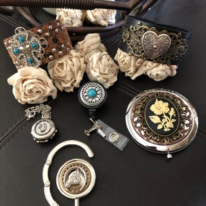 May include: A collection of western-themed jewellery and accessories, including a brown leather bracelet with turquoise stones, a black leather bracelet with a silver heart, a silver charm with a horseshoe, a silver charm with a horse head, and a silver compact mirror with a floral design.