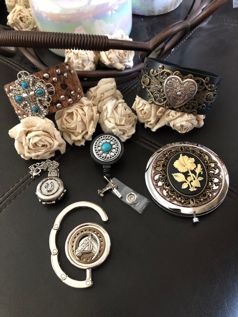 May include: A collection of western-themed jewellery and accessories, including a brown leather bracelet with turquoise stones, a black leather bracelet with a silver heart, a silver charm with a horseshoe, a silver charm with a horse head, and a silver compact mirror with a floral design.