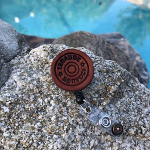 May include: A brown leather 12 gauge shotgun shell keychain with a black retractable reel. The keychain is on a gray and white rock surface.