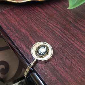 May include: A silver-toned metal purse hook featuring a round top with a decorative antique bronze-colored frame. Inside the frame is a black oval cameo inlay displaying a white floral design. The hook has a hinged arm for hanging bags.