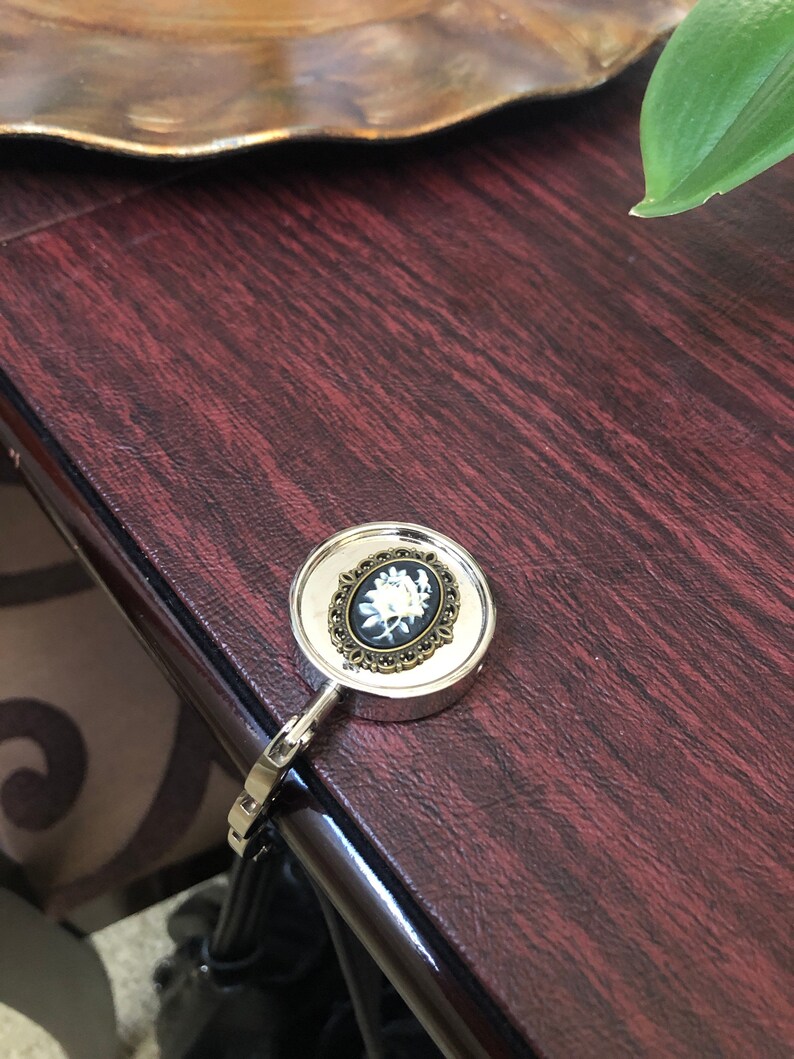 May include: A silver-toned metal purse hook featuring a round top with a decorative antique bronze-colored frame. Inside the frame is a black oval cameo inlay displaying a white floral design. The hook has a hinged arm for hanging bags.