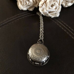 May include: A silver locket necklace with an ornate design. The locket has a circular shape and is decorated with a repeating pattern of circles and swirls. The necklace is made of a silver chain.