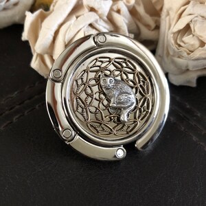 May include: Silver-toned metal purse hook with a decorative design featuring a cat sitting inside a circle of gold-toned metal.