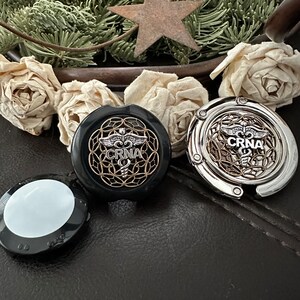 K&ouml;nnte beinhalten: Zwei silberne und schwarze Metall-Handtaschenhaken mit einem medizinischen Symbol und dem Text "CRNA" darauf. Die Haken befinden sich auf einer schwarzen Lederoberfl&auml;che.