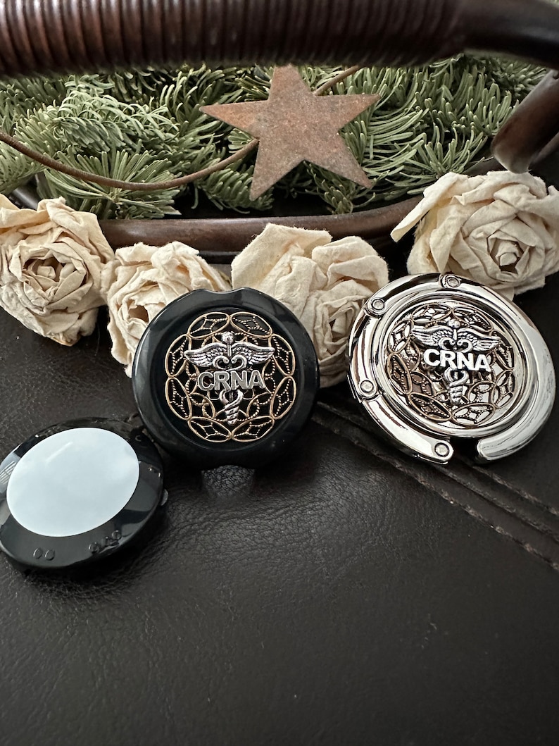 K&ouml;nnte beinhalten: Zwei silberne und schwarze Metall-Handtaschenhaken mit einem medizinischen Symbol und dem Text "CRNA" darauf. Die Haken befinden sich auf einer schwarzen Lederoberfl&auml;che.