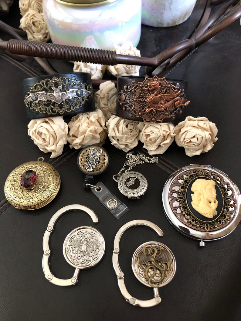 May include: A set of silver-toned metal clips with a bat and dragon design. The clips are attached to a black leather bracelet with a bat and dragon design. There is also a silver-toned metal locket with a bat design, a silver-toned metal locket with a dragon design, a silver-toned metal locket with an owl design, and a gold-toned metal locket with a cameo design.