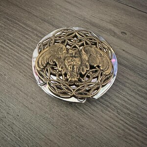 May include: A round, silver-toned compact mirror featuring an ornate, raised brass-colored metal overlay on the lid. The decorative design depicts a central owl with outstretched wings, surrounded by an intricate, woven, rope-like circular pattern.