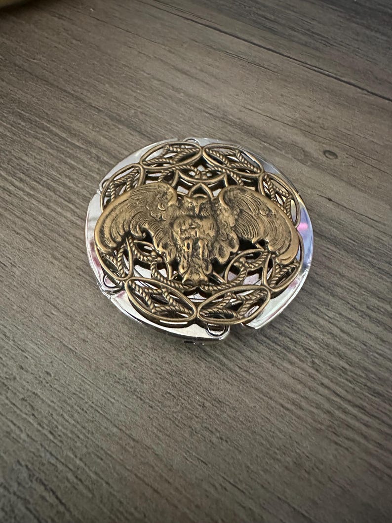 May include: A round, silver-toned compact mirror featuring an ornate, raised brass-colored metal overlay on the lid. The decorative design depicts a central owl with outstretched wings, surrounded by an intricate, woven, rope-like circular pattern.