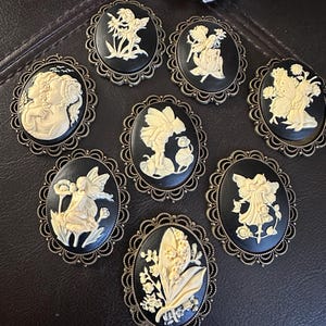 May include: A collection of seven oval brooches featuring cream-colored relief carvings on black backgrounds. Each piece is set in an ornate, scalloped bronze-toned metal frame. The designs depict various fairy figures and floral arrangements.