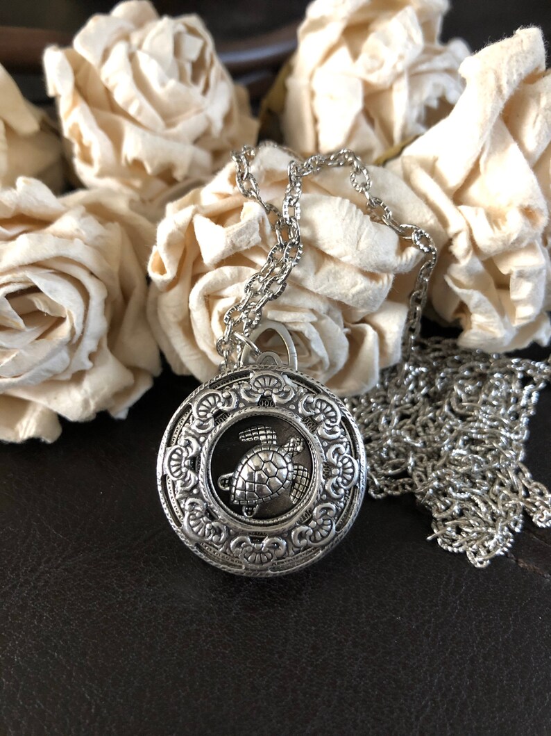 May include: A silver necklace with a round pendant featuring a detailed design of a sea turtle. The pendant is surrounded by a decorative border.