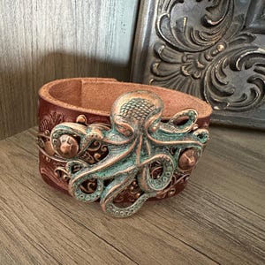 May include: A brown leather cuff bracelet with a copper and green octopus design. The octopus is detailed with textured scales and intricate swirls.