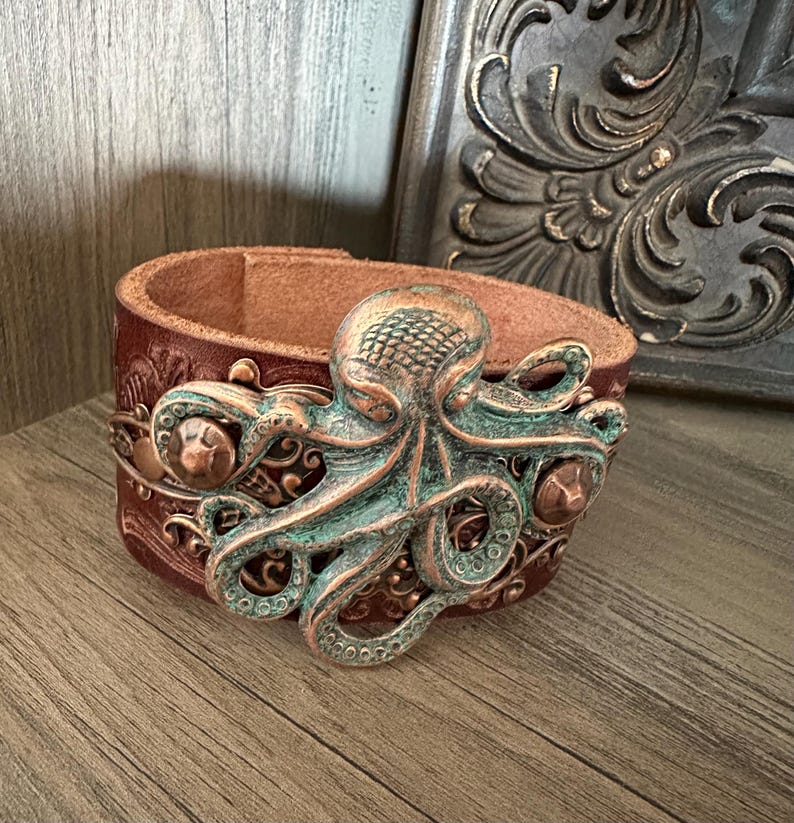May include: A brown leather cuff bracelet with a copper and green octopus design. The octopus is detailed with textured scales and intricate swirls.