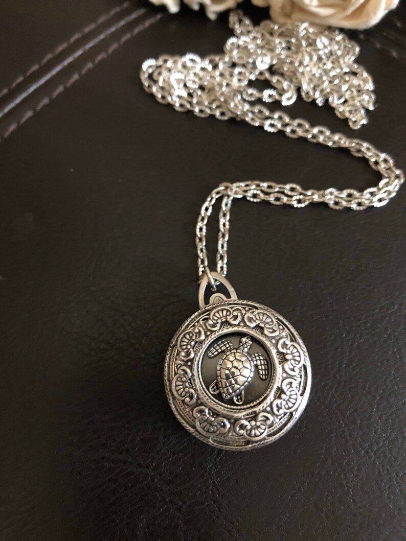 May include: A silver chain necklace with a round pendant featuring an ornate design and a silver turtle charm.