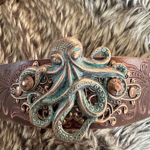May include: A brown leather belt with an ornate metal buckle featuring a green and copper octopus design.