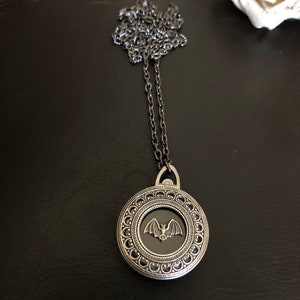May include: A silver necklace with a round pendant featuring a black enamel bat design. The pendant has a detailed silver frame with a beaded border.