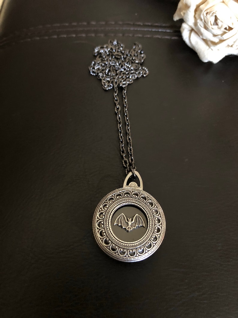 May include: A silver necklace with a round pendant featuring a black enamel bat design. The pendant has a detailed silver frame with a beaded border.