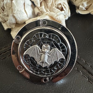 May include: A round, silver-toned metal purse hook featuring a central circular inlay. The inlay displays a detailed, raised silver bat figure positioned over a black, intricate web-like pattern. The outer edge of the hook has a smooth, polished finish.