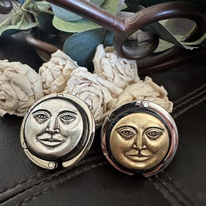May include: Two round, antique-style brooches with a smiling moon face design. One brooch is silver-toned, the other is gold-toned. Both have a black border and a pin back. The brooches are displayed on a dark brown surface.