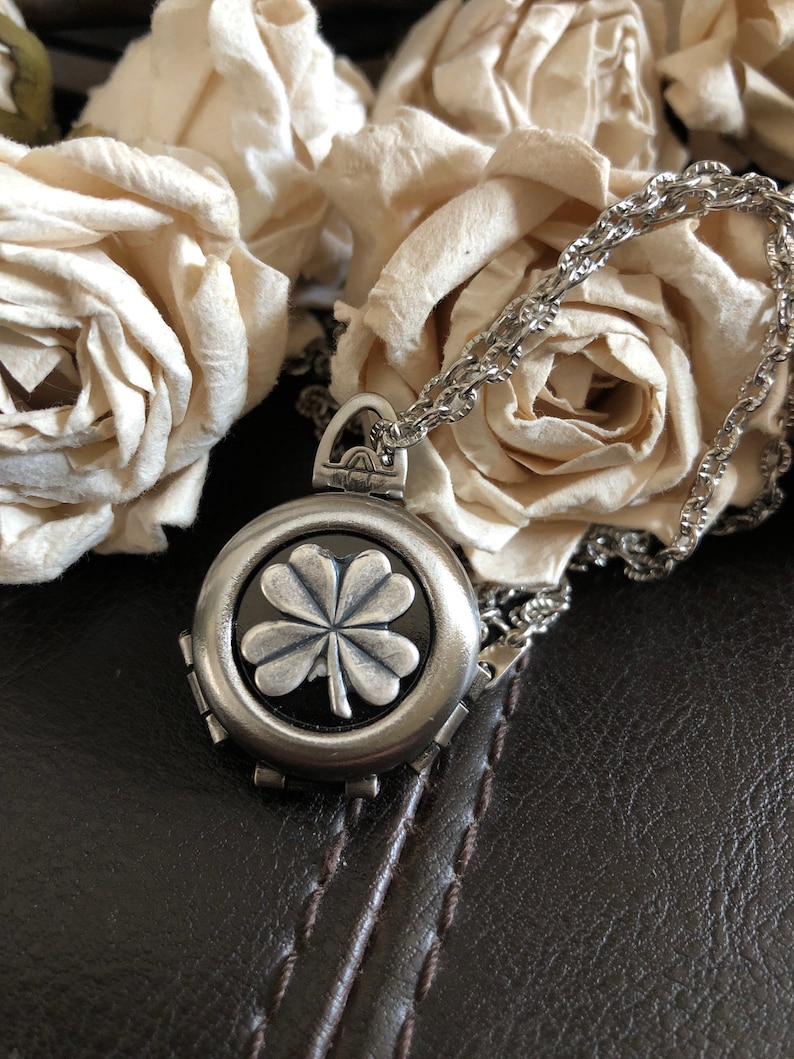 May include: A silver necklace with a four-leaf clover charm pendant. The pendant is round and has a black background. The necklace is on a brown leather surface.