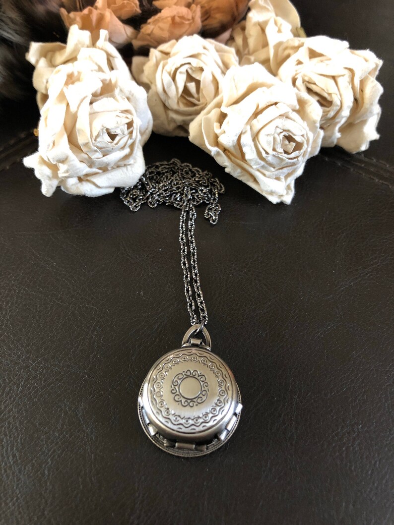 May include: A silver locket necklace with an ornate design. The locket is round and has a detailed pattern around the edge. The chain is silver and has a delicate design.