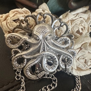 May include: A silver-toned octopus-shaped belt buckle with intricate detailing. The octopus has a textured head and curling tentacles. A silver chain is attached to the buckle. The buckle is set against a backdrop of cream-colored fabric.