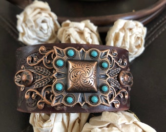 May include: A wide, brown leather cuff bracelet with a decorative copper-colored metal centerpiece. The centerpiece features a square design with turquoise-colored stones and intricate detailing. The bracelet is surrounded by cream-colored fabric flowers.