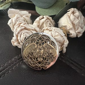 May include: A round, silver-toned metal compact mirror featuring an intricate, raised brass-colored owl emblem at its center. The owl is depicted with wings spread wide, surrounded by a decorative, openwork filigree pattern within the circular frame.