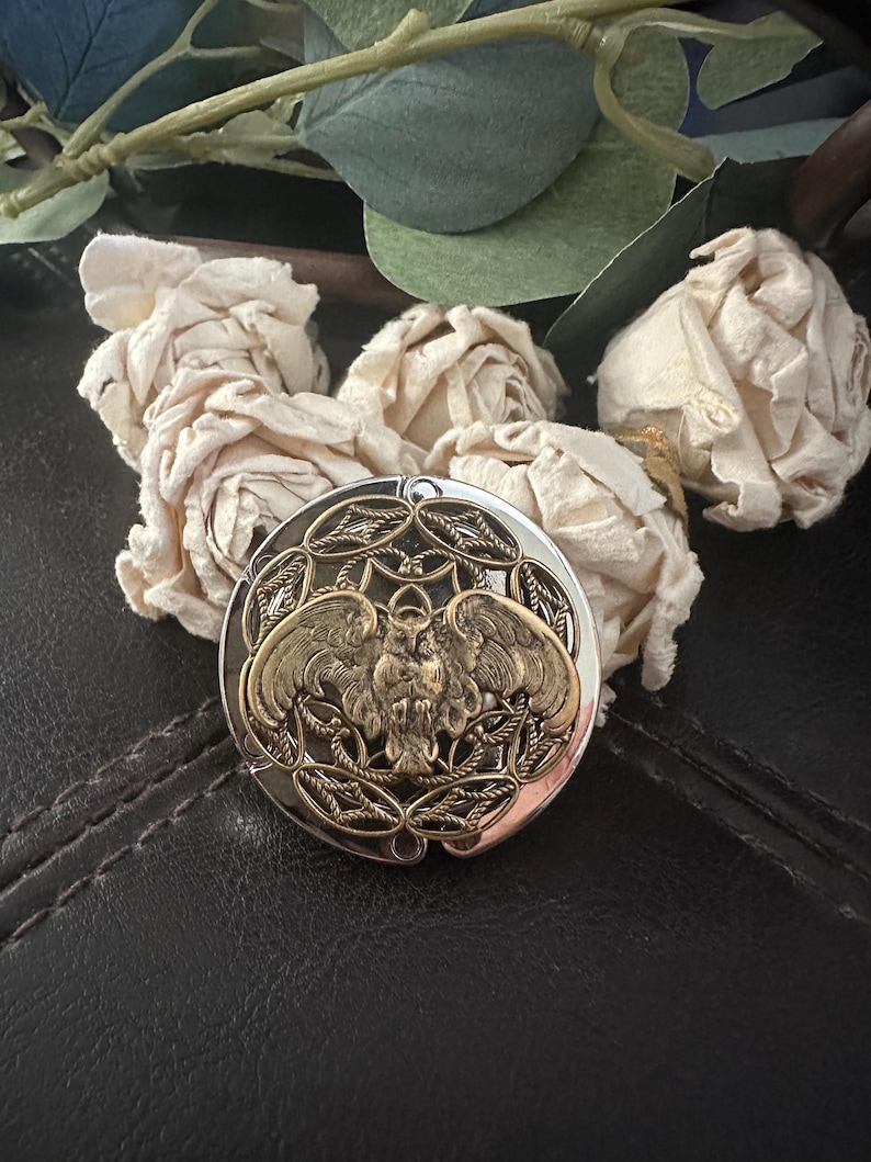 May include: A round, silver-toned metal compact mirror featuring an intricate, raised brass-colored owl emblem at its center. The owl is depicted with wings spread wide, surrounded by a decorative, openwork filigree pattern within the circular frame.