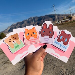 以下が含まれることがあります： 手には、漫画の動物のデザインが施された4つのマグネットブックマークがあります。各ブックマークには、犬、猫、ウサギ、クマなど、それぞれ悲しい表情と駄洒落に基づいたフレーズが特徴です。ブックマークはピンク色で、白い雲のアクセントが付いています。