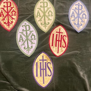 May include: Six oval-shaped embroidered patches featuring religious symbols. Four patches display the Chi-Rho monogram with Alpha and Omega letters in red, light green, light blue, and lavender. Two patches feature the IHS monogram in red and purple.