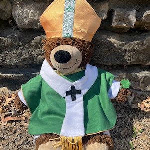 Puede incluir: Un oso de peluche marrón con una mitra dorada y verde y una casulla verde con cuello blanco y cruz negra. El oso tiene un trébol adjunto a su brazo.