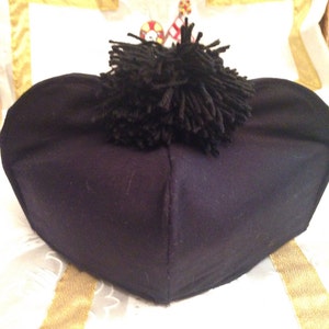 May include: A black fabric cap with a black pom-pom on top. The cap is folded in half and is sitting on a white surface with gold trim.