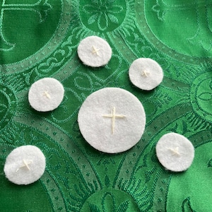 May include: Five white communion wafers with embroidered crosses on a green fabric background.